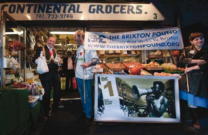 Two men stand outside a grocer's, one holding a bag, the other next to a large banner promoting "The Brixton Pound" local currency.