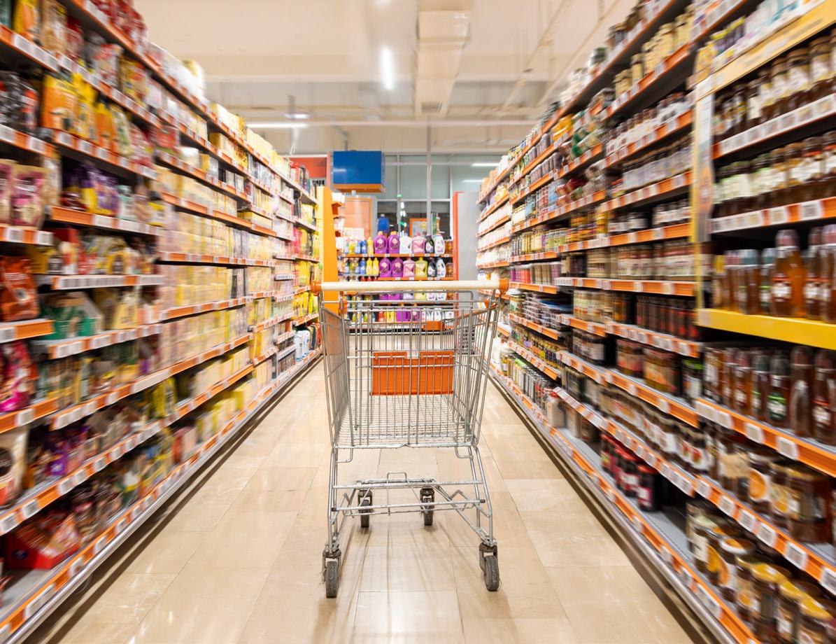 A wide perspective shot down a supermarket aisle with a shopping cart centered, flanked by long shelves stocked with various products.