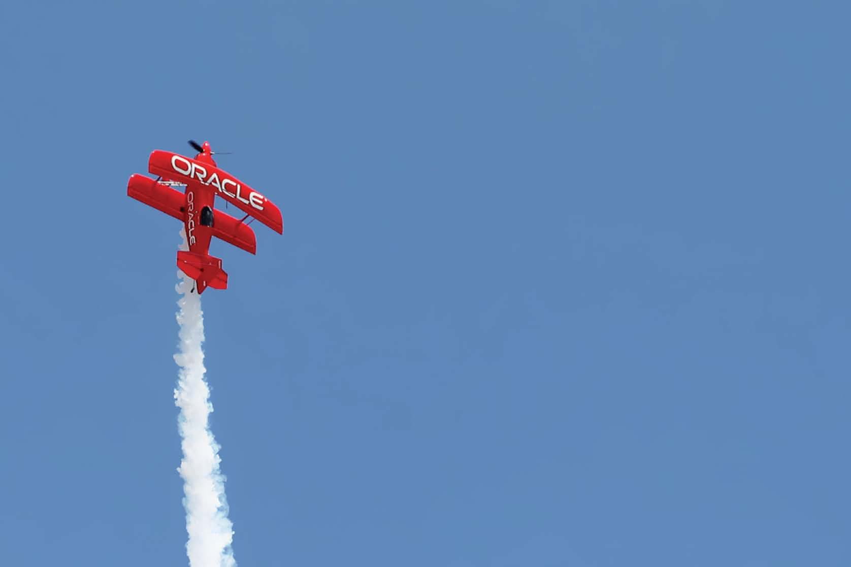A red Oracle branded biplane performs an aerial maneuver, leaving a white smoke trail against a clear blue sky.