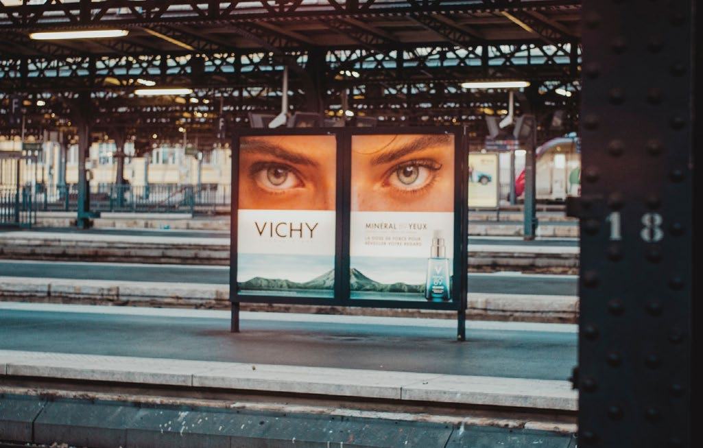 Advertisement billboard featuring large eyes and the brand 'VICHY' on a train station platform.