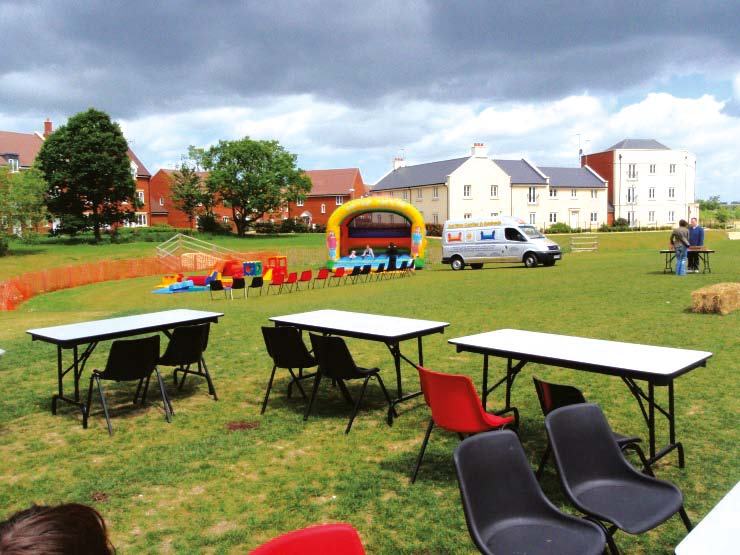 An outdoor community event on a grassy field with residential buildings, featuring a bouncy castle, white van, tables, chairs, and hay bales.