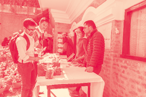 Several people interact around a table at an outdoor event or market stall, possibly signing up or receiving information.