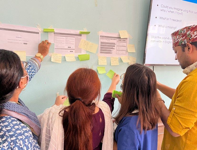 Four people participating in a workshop, placing sticky notes on papers attached to a wall, with a screen displaying questions in the background.