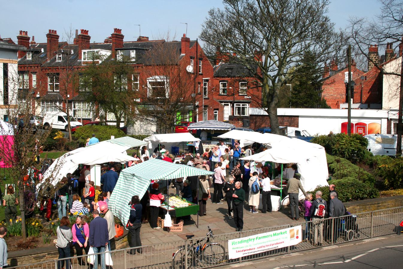 An outdoor farmers' market with numerous stalls and people walking around, with brick buildings in the background.