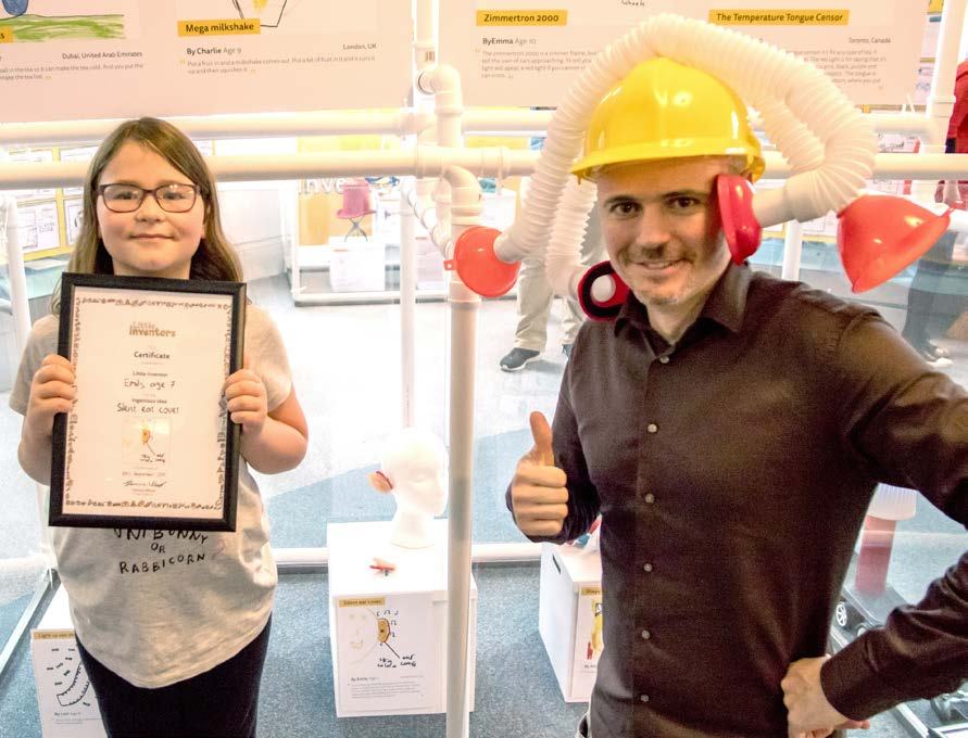 A girl holds a framed 'Junior Inventors' certificate next to an adult man in a yellow hard hat giving a thumbs-up.