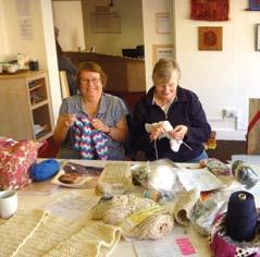Two women sitting at a table, smiling and engaged in knitting or crocheting, with yarn and craft supplies spread out before them.