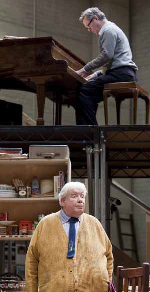 Two men on a multi-level stage set. An older man in a yellow cardigan stands below, looking out. Another man plays a piano on an elevated platform.