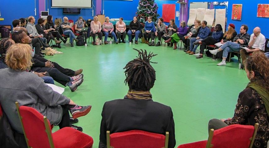 A diverse group of people sit in a large circle on chairs in a brightly lit room, engaged in a discussion or meeting.