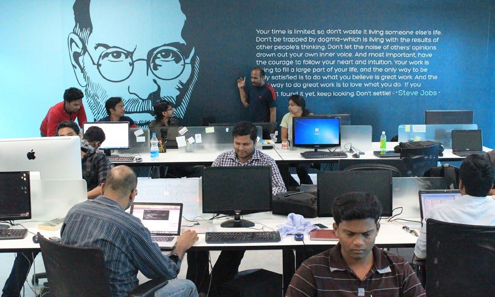 Employees working at desks in a modern office, with a large wall mural of Steve Jobs and an inspirational quote.