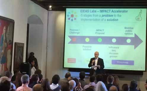 A man presents to an audience, with a large screen behind him displaying an "IDEAS Labs - IMPACT Accelerator" process diagram outlining 5 stages from problem to solution.