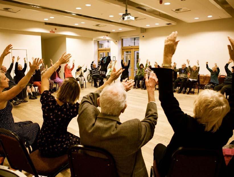 A group of mostly older adults raising their arms in unison during an activity in a large community hall.