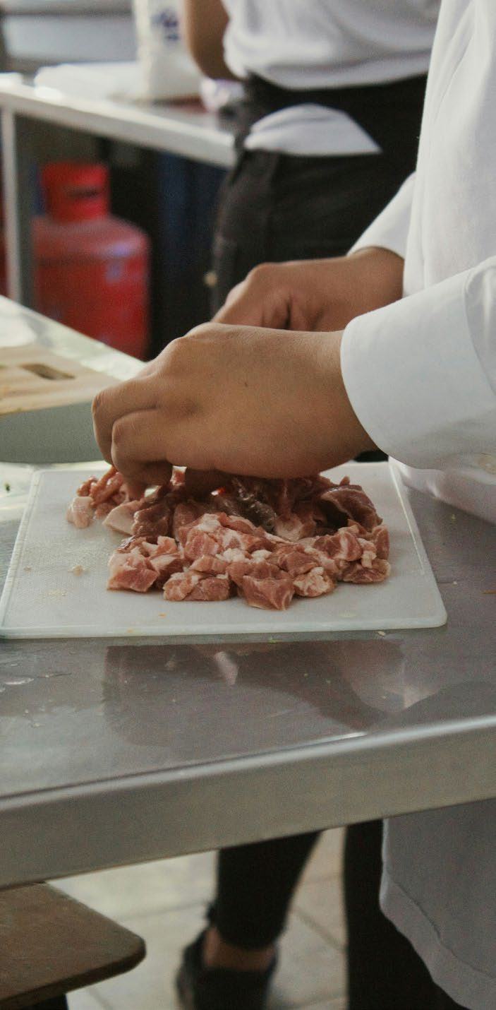 Close-up of hands in a white chef's coat, chopping raw meat into small pieces on a clear cutting board.