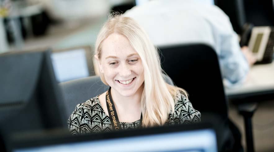 A young blonde woman smiling while working on a computer in a modern office environment.