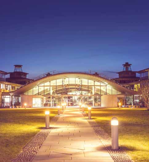 Exterior view of a modern building with a large curved glass facade, illuminated at night, with a paved walkway lined with glowing lights.