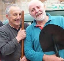 Two men smiling, one holding a wooden stick, the other a shovel, likely participating in an outdoor community project.