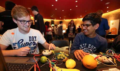 Two young men smiling, interacting with a laptop and various fruits connected by wires in an educational or experimental setting.