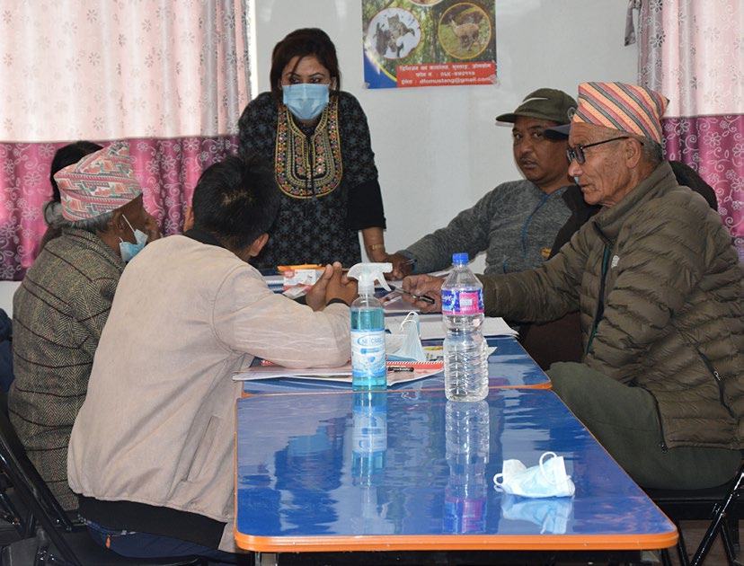 A group of people, some wearing masks, gathered around a table for a meeting or training session, with one woman standing.