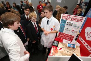 Students at a project booth featuring a display board titled "NHS in Distress," a model road network, and a tablet, with one student in a lab coat.