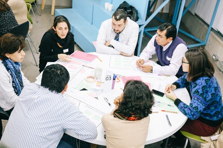 Group of people collaborating around a table covered with diagrams and papers during a workshop session.