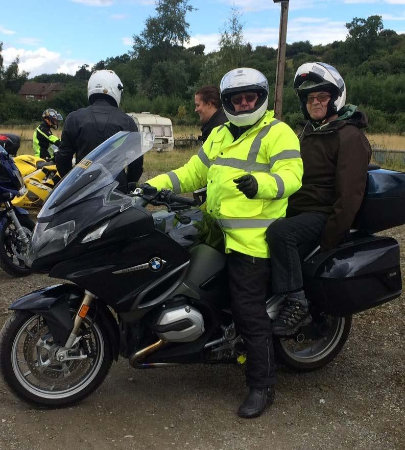 Two men in helmets on a black motorcycle, with other riders and bikes in the background, likely at an outdoor event.