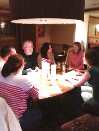 A group of people gathered around a large round wooden table in a dimly lit indoor setting, engaged in conversation.