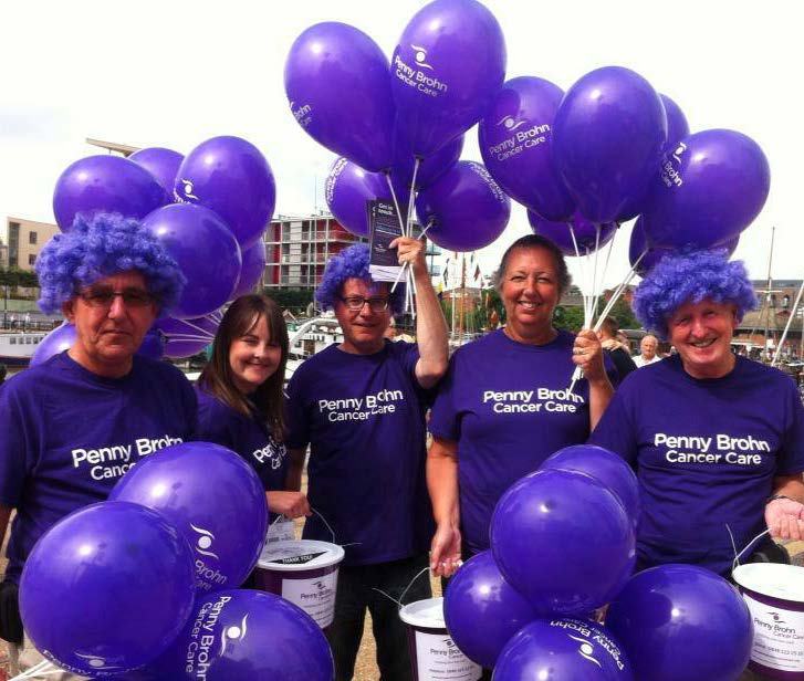 Five people in purple shirts and wigs, holding purple balloons and buckets for "Penny Brohn Cancer Care" fundraising event.