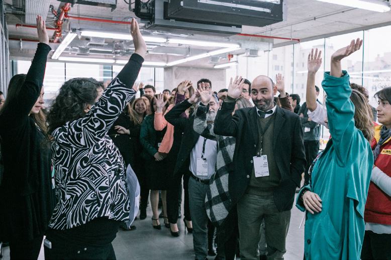 Diverse group of people in a modern office space enthusiastically raising their hands during an interactive session.