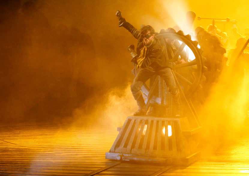 Actor in goggles and steampunk costume stands on a prop train with a large gear, amidst golden light and fog.