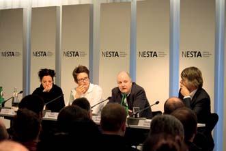 Panel discussion with four speakers at a table with microphones in front of a Nesta branded backdrop.