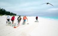 Group of people on a white sand beach pulling nets filled with marine debris, with a bird flying overhead and turquoise water in the background.