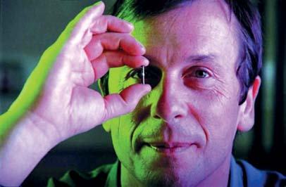 Man with green and purple lighting, holding a tiny microchip up to his eye, focusing intently.