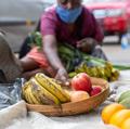 A person in a market stall reaching for bananas and apples in a wooden bowl, depicting local commerce.