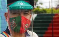 Person wearing a face shield and a red and green mask with unique symbols, looking forward.