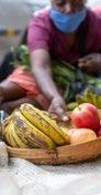 Close-up of bananas and apples in a wooden bowl, with a person in a mask reaching for items in the background.
