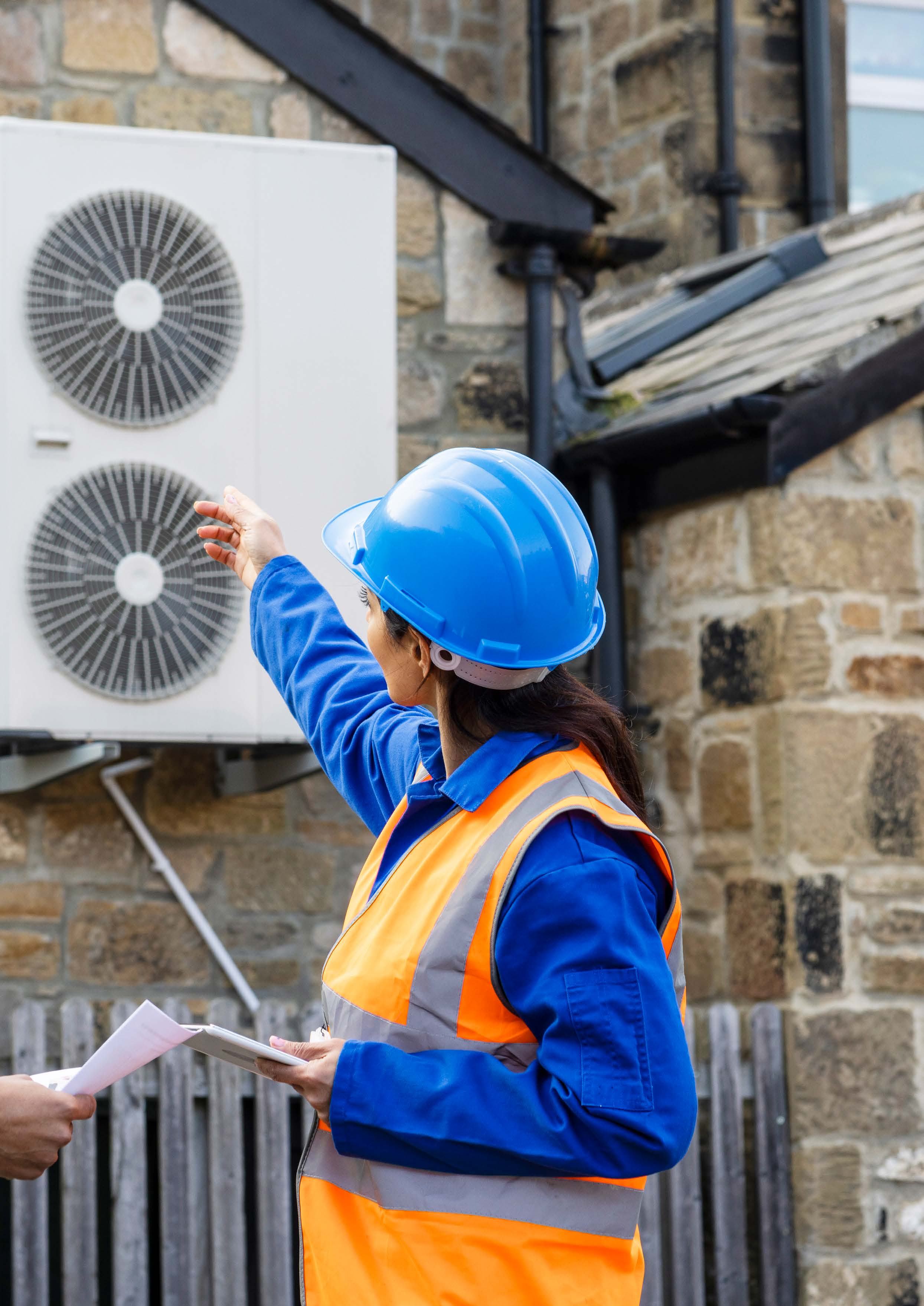 A worker in a hard hat and safety vest points at an outdoor air conditioning unit while discussing documents with another person.
