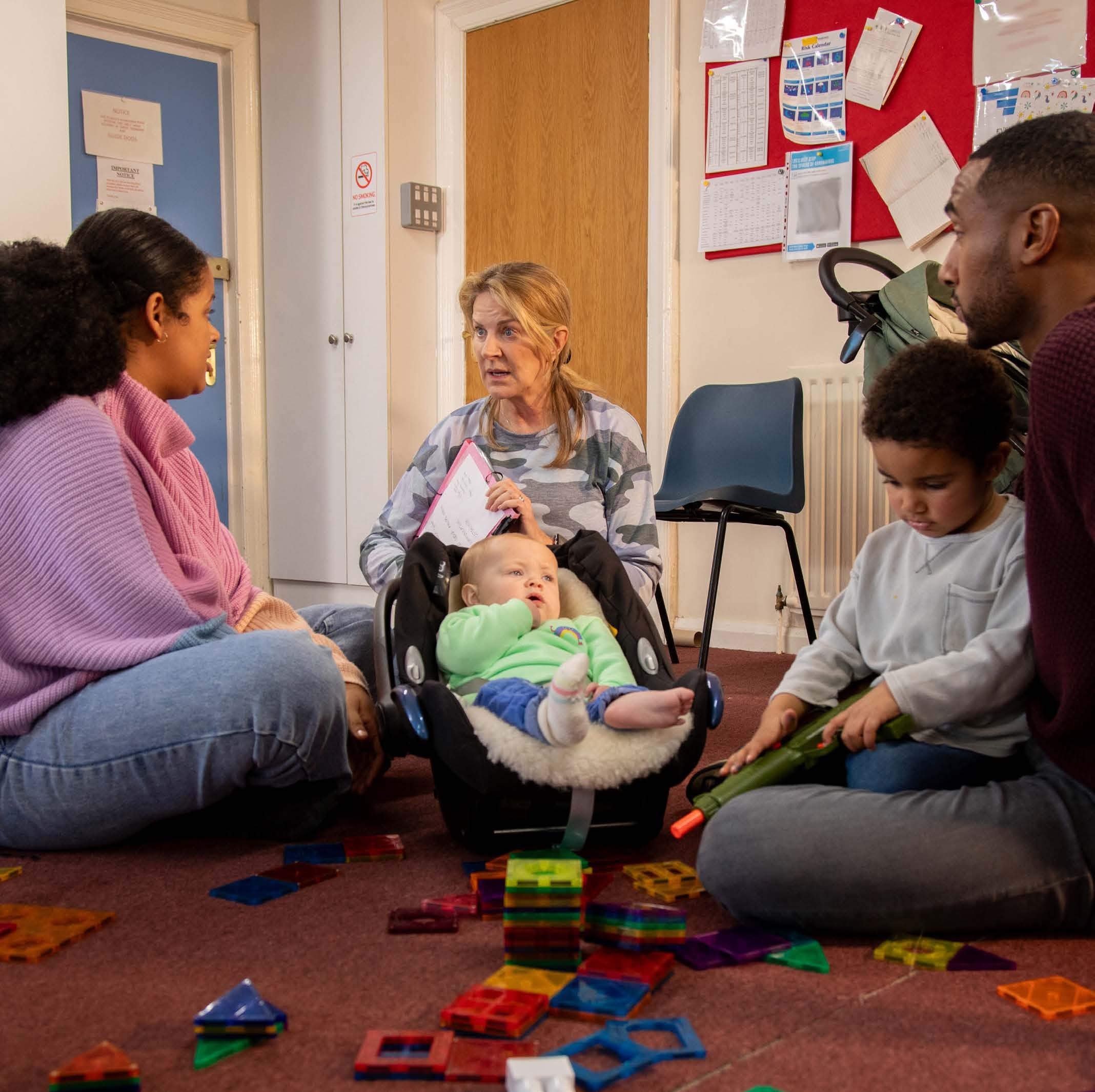 A diverse group of adults and children sitting on a carpeted floor in a room, interacting and playing with toys, possibly depicting a family support setting.