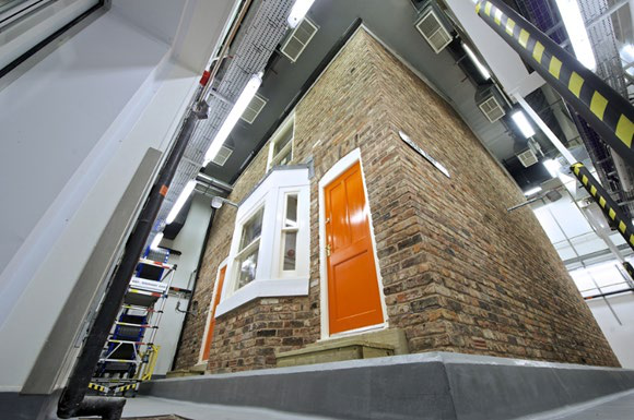 Interior view of the Energy House 2.0 lab, showing a brick terraced house test chamber with an orange door and industrial equipment for research.
