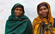 Two women wearing colorful headscarves, one green and one yellow, standing side-by-side and looking towards the camera.
