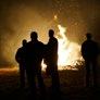 Silhouettes of people stand in front of a large, bright bonfire, casting long shadows in the dark