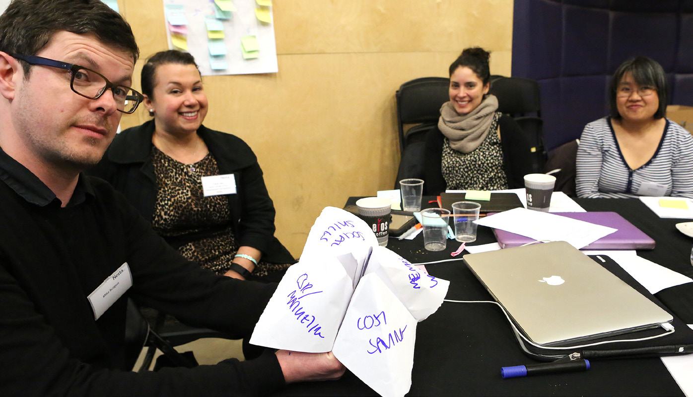 A group brainstorms with a man holding paper "fortune teller" objects labeled "Sales," "Marketing," and "Cost Saving."