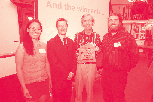 Four people stand on a stage, one holding an award, with a screen displaying "And the winner is..." in the background.