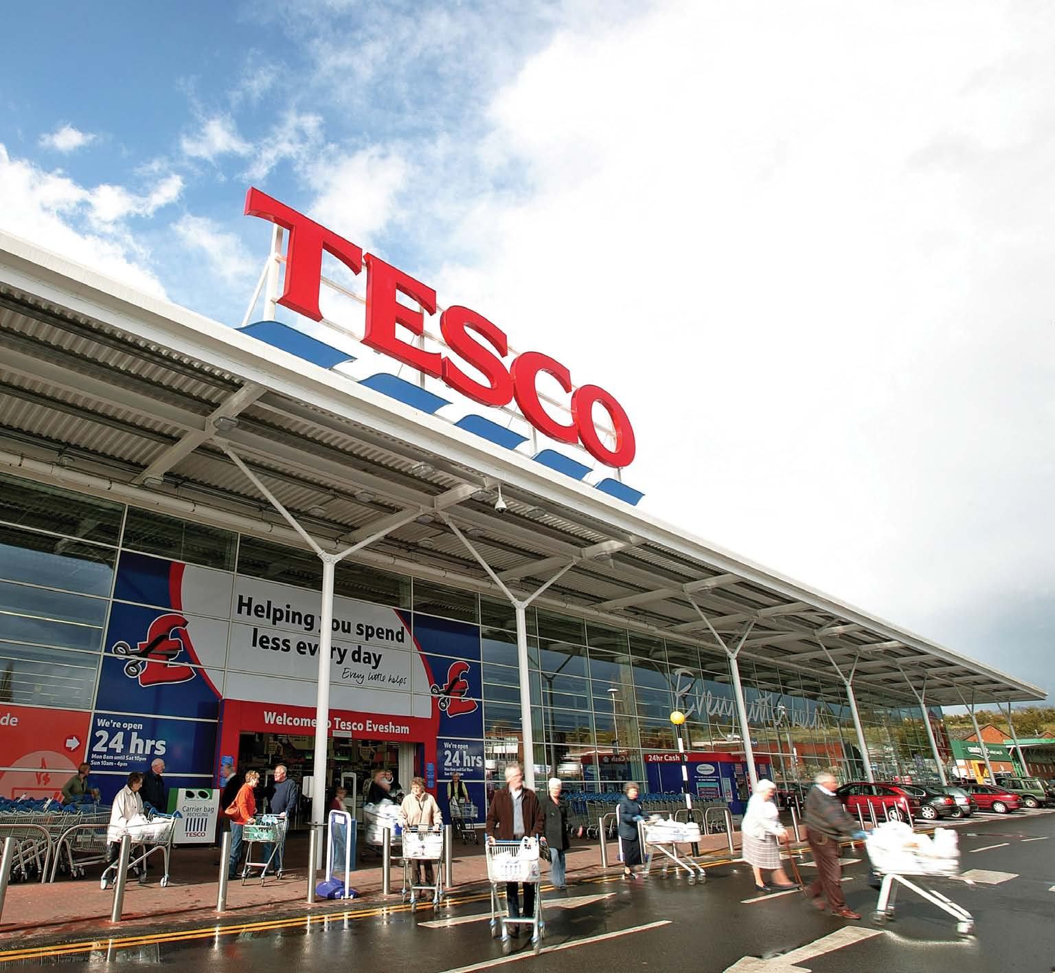 The exterior of a Tesco supermarket building with its prominent red logo, showing shoppers and cars under a cloudy sky.