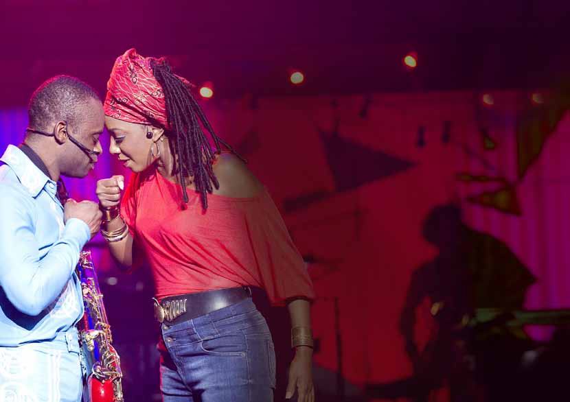 Two musicians on a stage with pink lighting. A man with a saxophone touches foreheads with a woman wearing a red top and red headwrap.