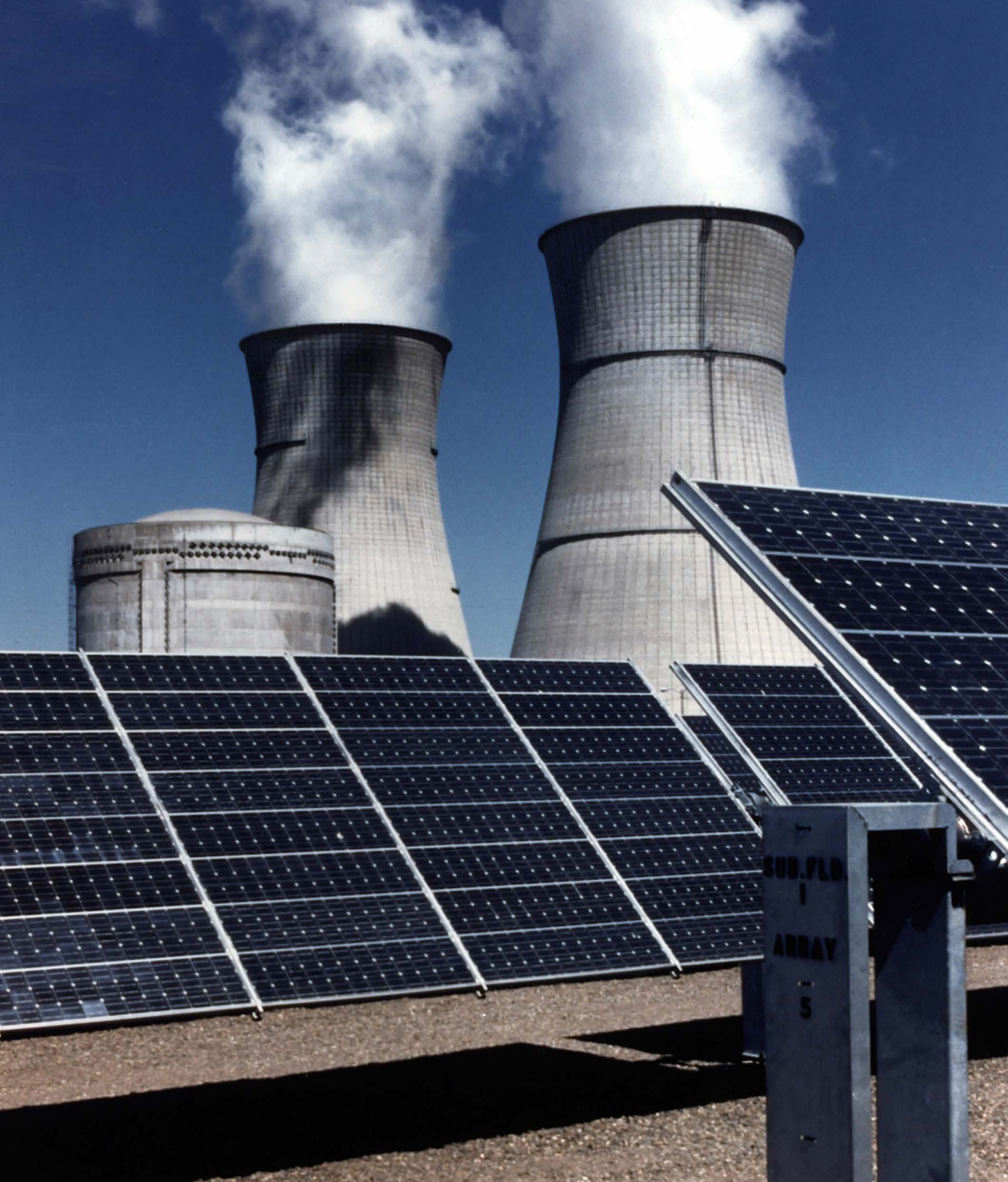 Solar panels in the foreground with large industrial cooling towers emitting steam in the background, contrasting renewable and traditional energy sources.