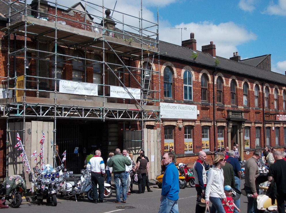 A street scene with an old brick building under scaffolding and people gathered with custom scooters on a sunny day.