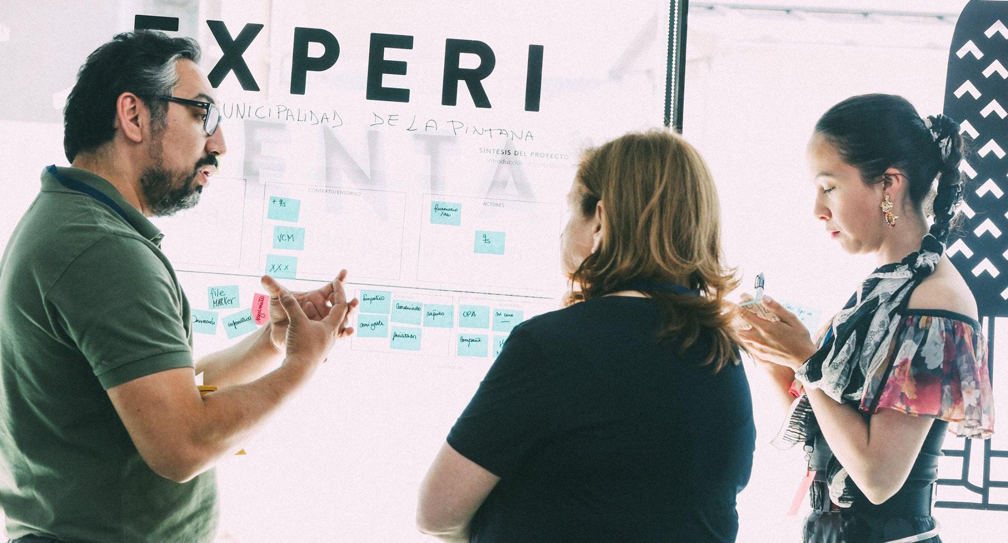 Three people discussing concepts written on a large transparent whiteboard, pointing at diagrams and text.