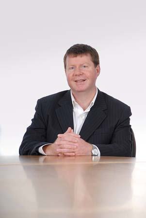 Portrait of a man in a dark suit and white shirt, sitting at a wooden table with hands clasped.