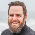 Headshot of a smiling man with a beard, wearing a dark turtleneck sweater, with a blurred ocean background.
