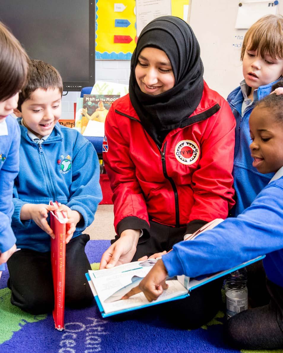 An adult woman in a red jacket and hijab reading a book with four young children on a colorful mat, illustrating an educational social action.