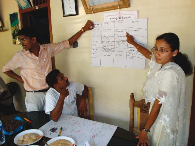 Three people discussing information, with a woman pointing at a handwritten chart on a wall and a man looking at a map on a table.